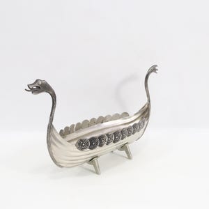 May include: A silver-colored Viking ship-shaped decorative bowl. The boat features a dragon head at the bow and stern, with a series of shield-like designs along the sides. The bowl is supported by three small legs and is set against a white background.