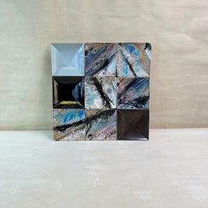 Könnte beinhalten: Neun quadratische Keramikfliesen, angeordnet in einem 3x3-Raster. Die Fliesen haben eine pyramidenförmige Form und eine Vielzahl von Farben und Mustern, darunter Blau, Schwarz, Braun und Weiß. Die Fliesen liegen auf einer hellen Oberfläche.
