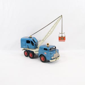 Pode incluir: Um brinquedo antigo de metal representando um caminhão-guindaste azul com um braço de elevação creme, um balde suspenso vermelho e rodas pretas com cubos vermelhos. A cabine tem uma grade frontal prateada, uma luz amarela e uma cabine azul.