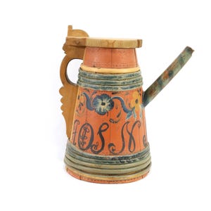 May include: A vintage, hand-painted wooden pitcher with a handle and spout. The pitcher features a vibrant orange body with blue floral and scroll designs, and the word "HOS" in black script. The handle and top are natural wood.