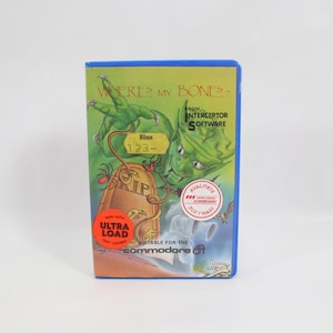 Puede incluir: Un cartucho de juego vintage de Commodore 64 titulado "Where's My Bones?". La carátula presenta un monstruo verde, una lápida y el texto "ULTRA LOAD". El cartucho tiene un marco azul y está sobre un fondo blanco.