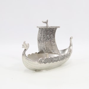 Op de afbeelding: Zilverkleurig Viking-scheepsbeeldje met het woord "NORGE" op het zeil. Het schip heeft decoratieve details en een drakenkop aan de voorkant. Het beeldje is gemaakt van metaal en staat op een witte ondergrond.