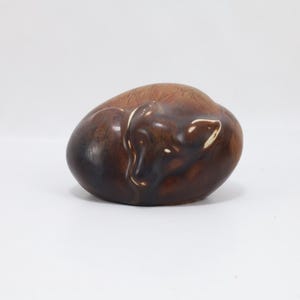 May include: A ceramic figurine of a sleeping fox, curled up in a ball. The fox is a rich brown color with lighter highlights, and the figurine has a glossy finish. The fox's ear is visible.