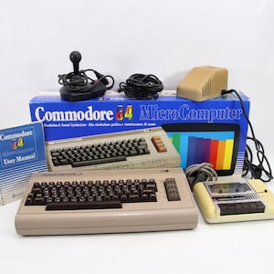 以下が含まれることがあります： 2 つのベージュ色のキーボード、ジョイスティック、カセットプレーヤー、電源を含む、ヴィンテージの Commodore 64 Microcomputer セット。 青い箱には Commodore 64 のロゴが表示されています。 ユーザーマニュアルも含まれています。