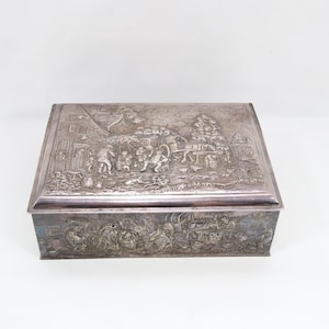 May include: A rectangular silver-toned box with detailed relief carvings. The lid and sides feature scenes of people, buildings, and animals. The box is set against a plain white background, highlighting its intricate design and antique aesthetic.
