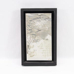 Op de afbeelding: Abstract kunstwerk in een zwarte rechthoekige lijst. Het kunstwerk toont een mix van grijze, witte en zwarte penseelstreken en texturen. De compositie bevat lagen verf en wat lijkt op tekstfragmenten.