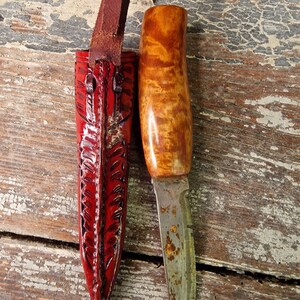 Puede incluir: Una peque&ntilde;a funda de cuchillo de cuero rojo con un cuchillo de mango de madera y una hoja oxidada.