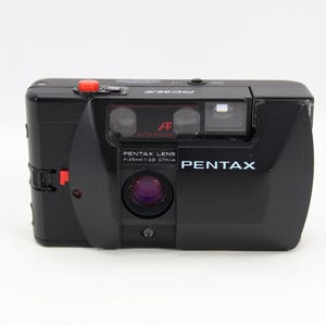 Może przedstawiać: Czarny aparat Pentax PC35AF z czerwonym przyciskiem migawki. Aparat posiada obiektyw z napisem "PENTAX LENS f=35mm 1:2.8 07m-∞" oraz nazwą marki "PENTAX" z przodu.