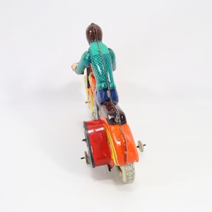 May include: A vintage toy motorcycle with a sidecar. The rider is wearing a green and blue outfit and a brown helmet. The motorcycle is painted in orange, yellow, and red with a red sidecar. The toy has a retro aesthetic.