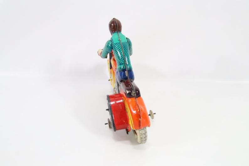 May include: A vintage toy motorcycle with a sidecar. The rider is wearing a green and blue outfit and a brown helmet. The motorcycle is painted in orange, yellow, and red with a red sidecar. The toy has a retro aesthetic.