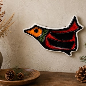 May include: A ceramic wall hanging in the shape of a bird, featuring a black body with red and green accents, and a white border. The bird is mounted on a beige wall. A vase with dried plants, a wooden bowl with pinecones, and a sprig of pine are also visible.