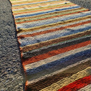 May include: A long, narrow, multi-colored striped rug with a woven texture. The rug is made of natural fibers and has a rustic, vintage look.