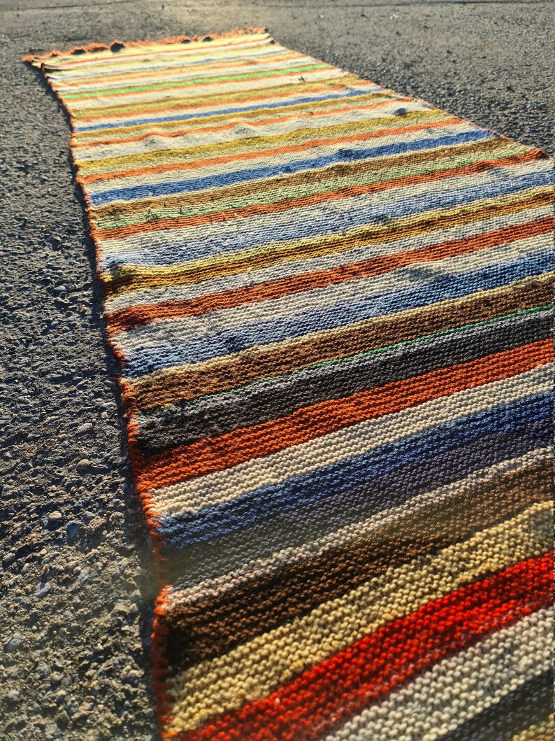 May include: A long, narrow, multi-colored striped rug with a woven texture. The rug is made of natural fibers and has a rustic, vintage look.