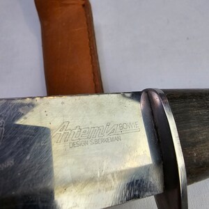 May include: A silver knife with a black wooden handle. The knife has the words "Artemis Bowie" and "Design S.Berkeman" engraved on the blade.