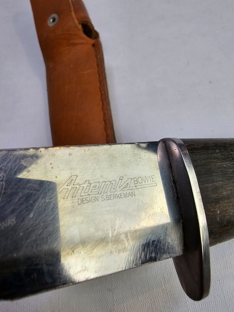 May include: A silver knife with a black wooden handle. The knife has the words "Artemis Bowie" and "Design S.Berkeman" engraved on the blade.