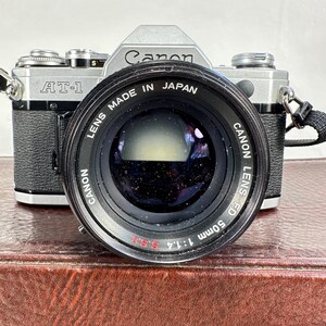 May include: A silver Canon AT-1 camera with a Canon FD 50mm 1:1.4 lens attached. The lens is black and has the text "Canon Lens FD 50mm 1:1.4 5.SS" printed on it.