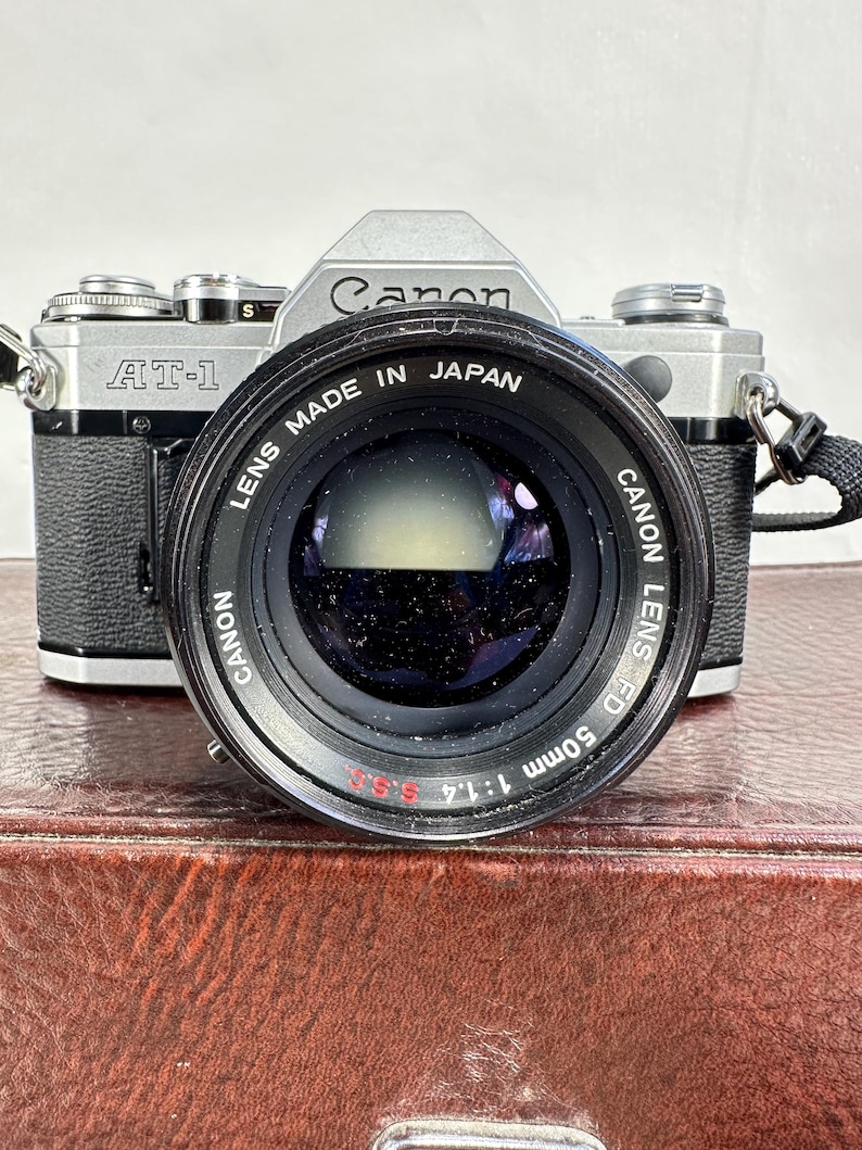 May include: A silver Canon AT-1 camera with a Canon FD 50mm 1:1.4 lens attached. The lens is black and has the text "Canon Lens FD 50mm 1:1.4 5.SS" printed on it.