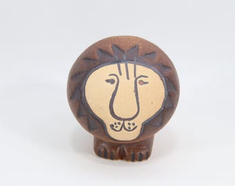 Może przedstawiać: Ceramiczna figurka lwa z brązową grzywą i ciałem oraz beżową twarzą. Twarz lwa ma czarne kontury oczu, nosa i ust. Figurka jest przedmiotem dekoracyjnym.