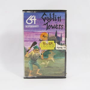 Puede incluir: Un cartucho de videojuego Goblin Towers de 64 Supersoft. La portada presenta un duende de dibujos animados con un garrote, un castillo y el texto "Goblin Towers". La cinta tiene una etiqueta de precio.