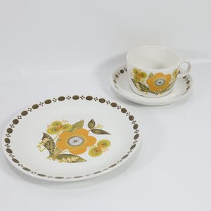 May include: A white ceramic plate, saucer, and teacup set with a floral design in yellow, orange, and green. The plate has a brown dotted border. The teacup and saucer also feature the floral pattern.