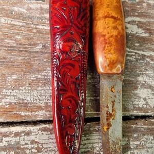 Puede incluir: Una funda de cuchillo de cuero rojo con intrincados tallados florales y un cuchillo de mango de madera con una hoja oxidada.