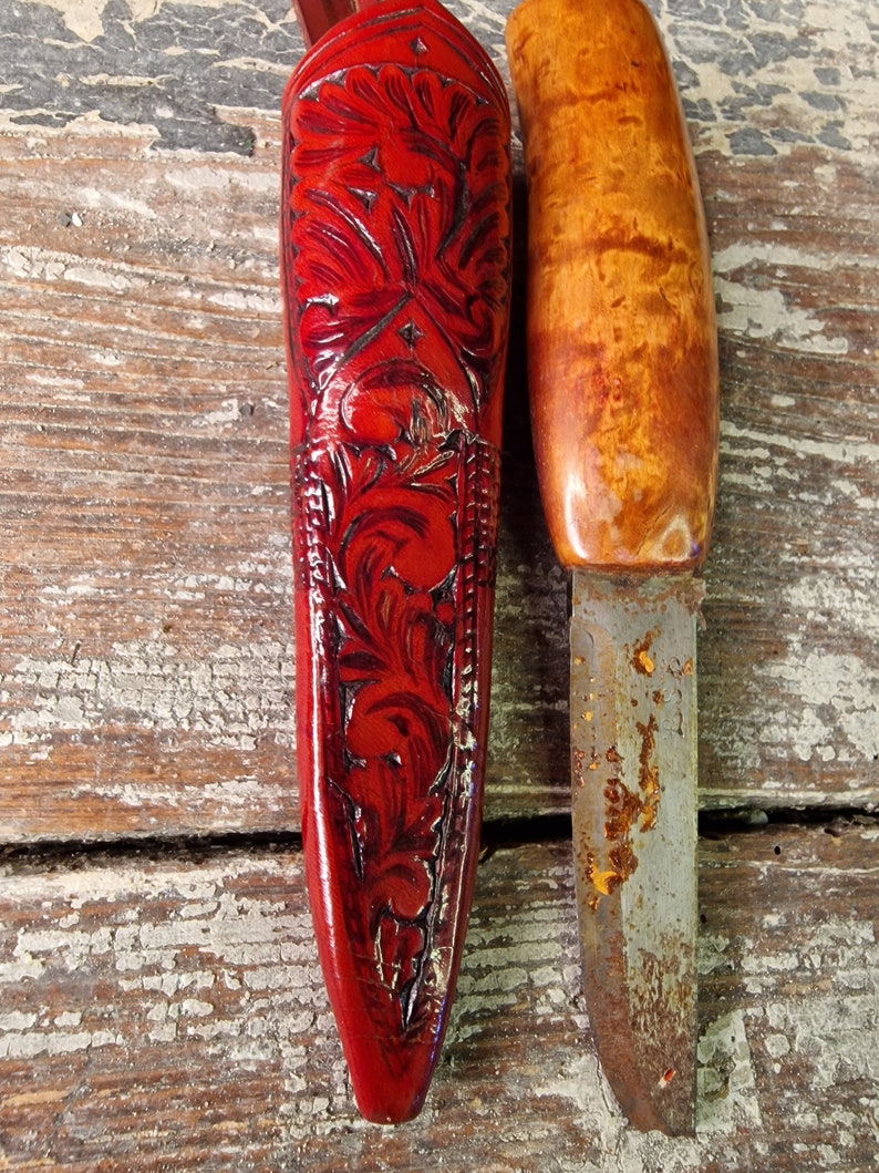 Puede incluir: Una funda de cuchillo de cuero rojo con intrincados tallados florales y un cuchillo de mango de madera con una hoja oxidada.
