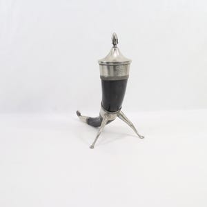 Op de afbeelding: Een donkere, gebogen drinkhoorn met een gepolijste zilverkleurige metalen rand, een puntig deksel met een kleine knop en een metalen standaard met drie poten. De zilverkleurige metalen band aan de bovenzijde bevat de gravure G.R.L.