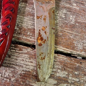Puede incluir: Primer plano de una hoja de cuchillo oxidada con mango de madera. La hoja est&aacute; parcialmente cubierta de &oacute;xido y tiene una ligera curva. El mango es de madera y tiene un acabado natural.