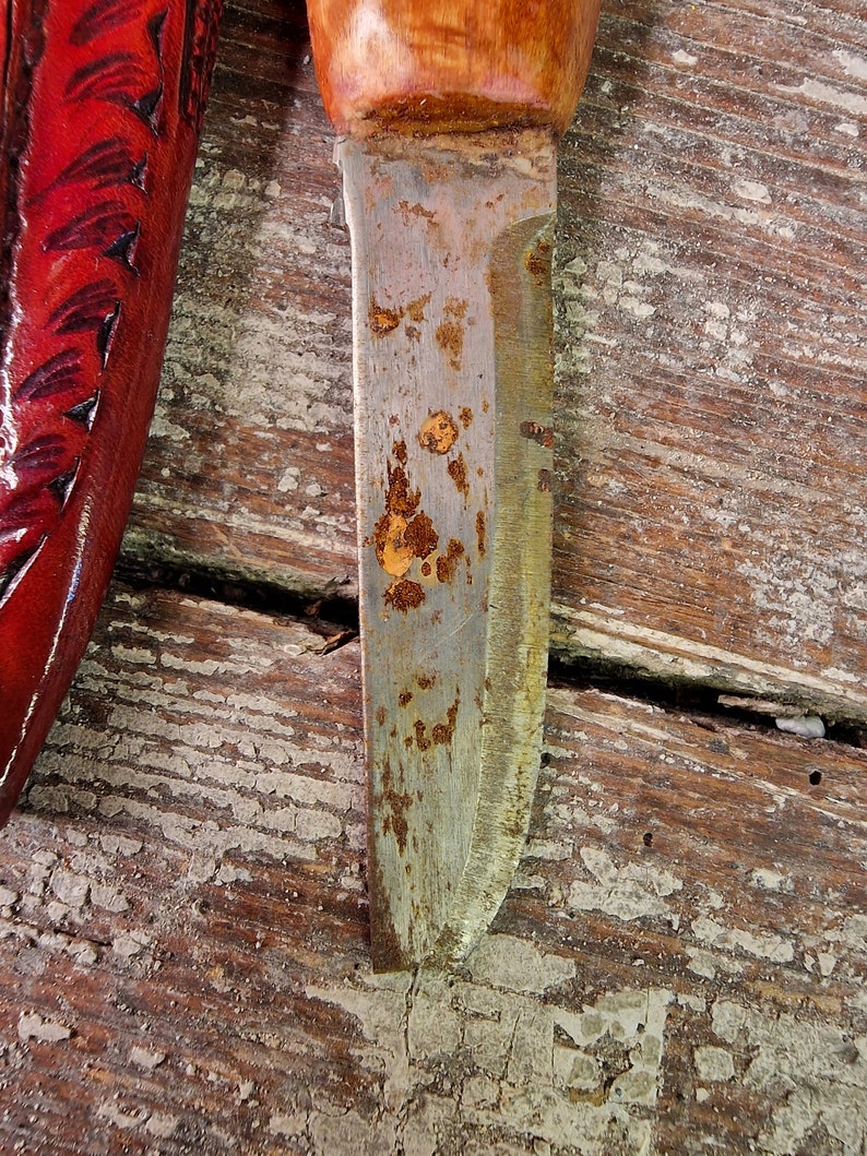 Puede incluir: Primer plano de una hoja de cuchillo oxidada con mango de madera. La hoja est&aacute; parcialmente cubierta de &oacute;xido y tiene una ligera curva. El mango es de madera y tiene un acabado natural.