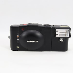 Puede incluir: Cámara Olympus DX negra con flash incorporado y el texto "OLYMPUS" en la parte delantera. La cámara tiene una lente redondeada y un pequeño flash en el lado derecho. El número de modelo "A11" es visible.