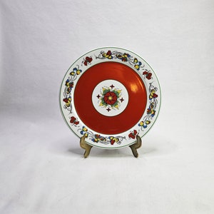 May include: A white ceramic plate with a red centre and a green, yellow, and blue floral design around the edge. The plate is on a gold metal stand.