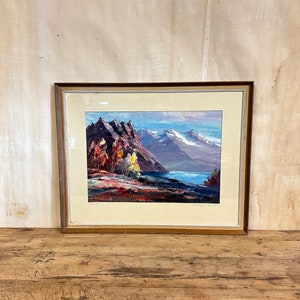 May include: A framed painting depicting a mountain landscape with a lake in the foreground. The mountains are painted in shades of brown, purple, and white, and the lake is a deep blue. There are trees in the foreground, painted in shades of green, yellow, and orange.