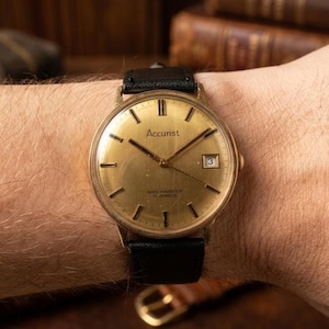 May include: A vintage Accurist wristwatch with a round gold-tone dial, black hour markers, and a date window at the three o'clock position. The watch face features the text Accurist, Shockmaster, and 17 Jewels, paired with a black leather strap.