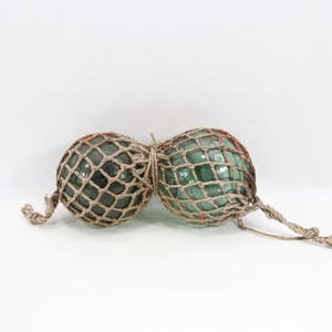 Puede incluir: Dos flotadores de pesca de vidrio vintage, cada uno envuelto en una red anudada. Los flotadores son de color verde mar y la red es de color beige natural. Los flotadores están atados con una cuerda natural.