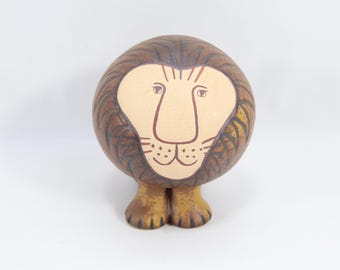 May include: A ceramic lion ornament with a rounded body and short legs. The lion's face is light beige with brown facial features. The mane is brown with dark brown stripes. The legs are a light brown colour.