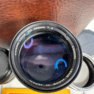 May include: A black Canon FD 200mm 1:4 lens with the text "Canon Lens FD 200mm 1:4 Canon Lens Made in Japan" printed on the side.