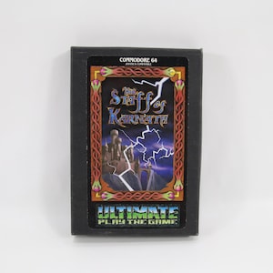 Puede incluir: Cartucho de juego Commodore 64 para "The Staff of Karnath". La caja negra tiene una etiqueta colorida con un castillo, relámpagos y el título del juego. El texto "ULTIMATE PLAY THE GAME" está en la parte inferior.