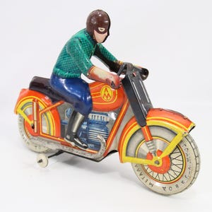 May include: Vintage toy motorcycle with a rider. The motorcycle is primarily orange with yellow and silver accents. The rider wears a green shirt, blue pants, and a brown helmet. The toy has a wind-up mechanism.