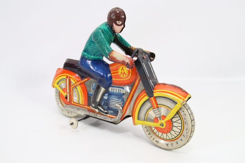 May include: Vintage toy motorcycle with a rider. The motorcycle is primarily orange with yellow and silver accents. The rider wears a green shirt, blue pants, and a brown helmet. The toy has a wind-up mechanism.