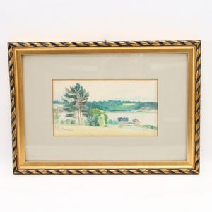 Könnte beinhalten: Ein gerahmtes Aquarell-Landschaftsbild mit einer ruhigen Seeufer-Szene mit Bäumen, einem Haus und einem Gewässer. Das Gemälde befindet sich in einem goldfarbenen Rahmen mit einem seilartigen Design.