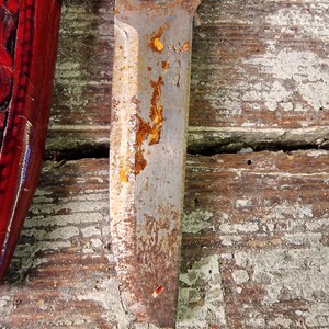 Puede incluir: Primer plano de una hoja de cuchillo oxidada con mango de madera. La hoja tiene algunas manchas de &oacute;xido y un peque&ntilde;o agujero cerca de la punta. La hoja est&aacute; apoyada sobre una superficie de madera.