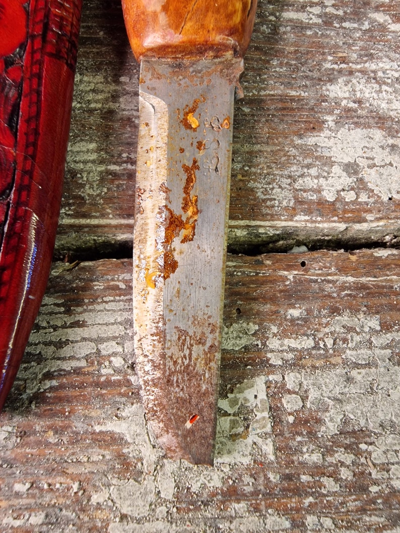 Puede incluir: Primer plano de una hoja de cuchillo oxidada con mango de madera. La hoja tiene algunas manchas de &oacute;xido y un peque&ntilde;o agujero cerca de la punta. La hoja est&aacute; apoyada sobre una superficie de madera.