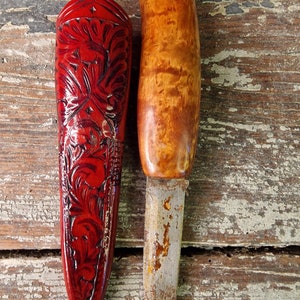 Puede incluir: Una funda de cuero roja con un intrincado dise&ntilde;o floral y un cuchillo con mango de madera y hoja de metal.