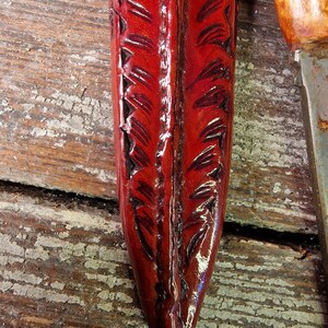 Puede incluir: Una funda de cuero roja con un patr&oacute;n decorativo de plumas. La funda est&aacute; hecha de cuero de alta calidad y tiene un acabado liso.