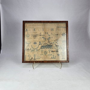 以下が含まれることがあります： クリーム色の背景に青と黒の刺繍が施された額装されたサンプラー。サンプラーには教会、木々、アルファベットの文字が描かれています。下部に「KT. BR. 1834」という文字が刺繍されています。