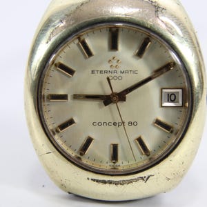 Puede incluir: Un reloj de pulsera vintage Eterna-Matic 3000 Concept 80. El reloj tiene una caja dorada y una esfera clara con marcadores y agujas negras. La fecha se muestra a las 3 en punto. Un reloj clásico.