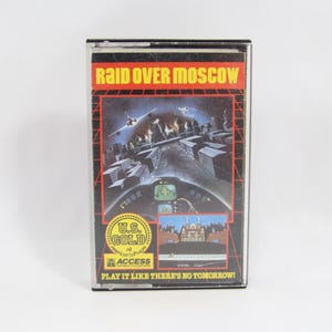 Puede incluir: Un cartucho de videojuego vintage titulado "Raid Over Moscow". La carátula presenta una ciudad futurista con aviones de combate y el título del juego en letras rojas. También es visible el logotipo de U.S. Gold. El texto "Play it like there's no tomorrow!" está impreso en la parte inferior.