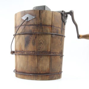 Può includere: Una gelatiera vintage in legno con una manovella manuale. Il secchio di legno è marrone e ha delle fasce metalliche. La manovella è in metallo e ha un manico in legno.