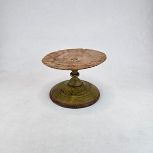 Antique wooden pedestal cake stand with round top and turned base, decorated with worn green and red painted surface. The top plate shows heavy age patina and visible wood grain with an aged central mark. The stem has carved details and a turned profile. Base is circular with layered rings and traces of original paint. Rustic primitive Scandinavian folk art stand photographed on white background.