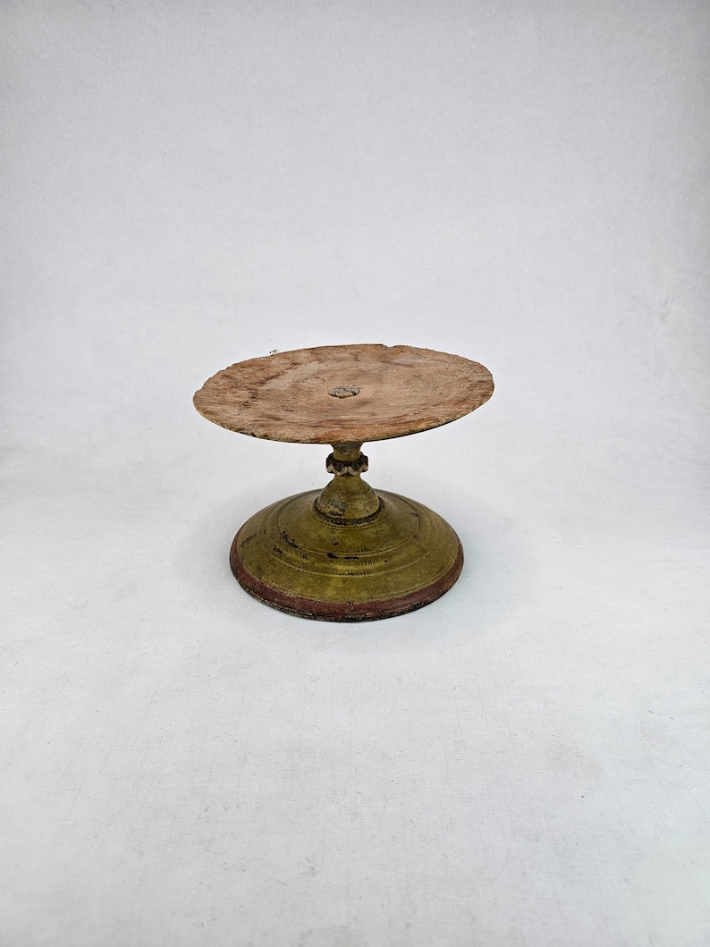 Antique wooden pedestal cake stand with round top and turned base, decorated with worn green and red painted surface. The top plate shows heavy age patina and visible wood grain with an aged central mark. The stem has carved details and a turned profile. Base is circular with layered rings and traces of original paint. Rustic primitive Scandinavian folk art stand photographed on white background.